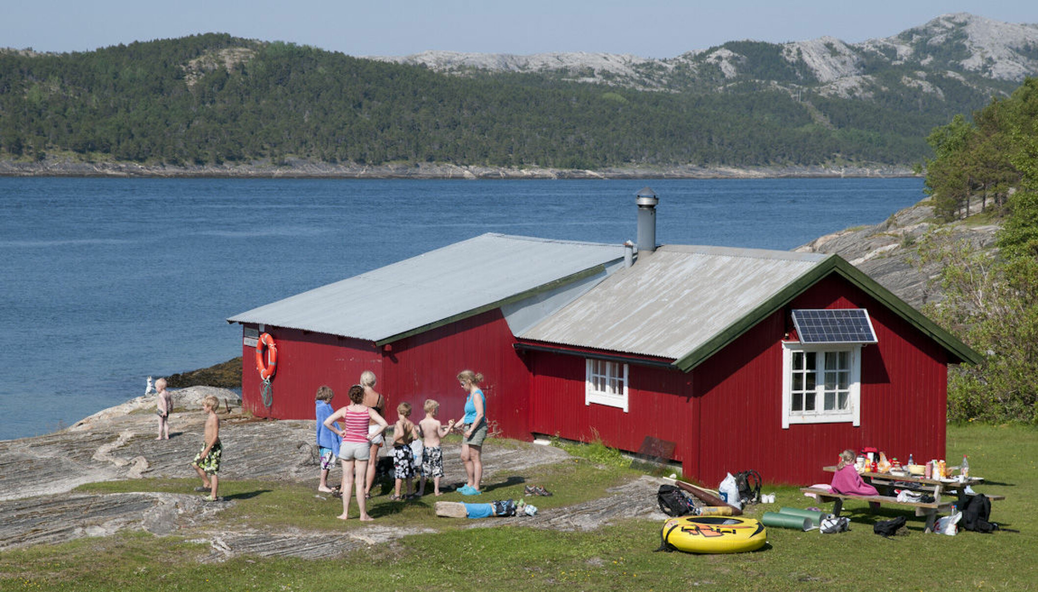 Flere voksne og barn leker og bader utenfor en rød hytte som ligger rett ved sjøen. 