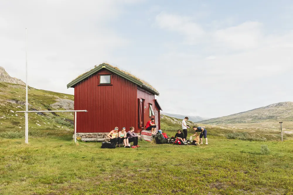 Rød hytte med grønt gress rundt. Personer som slapper av foran hytta.
