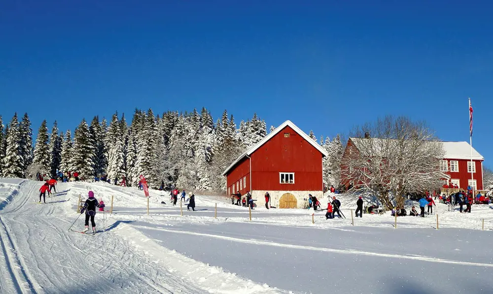 En stor rød hytte og en rød låve i utkanten av en skog, vinter og masse snø på trærne. Mennesker går i oppkjørte skispor foran hytta.
