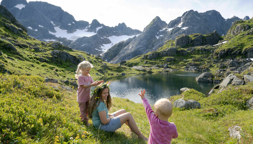 Kvinne med blomsterkrans sitter i lyngen i ei fjellside mens to små barn leker rundt henne.