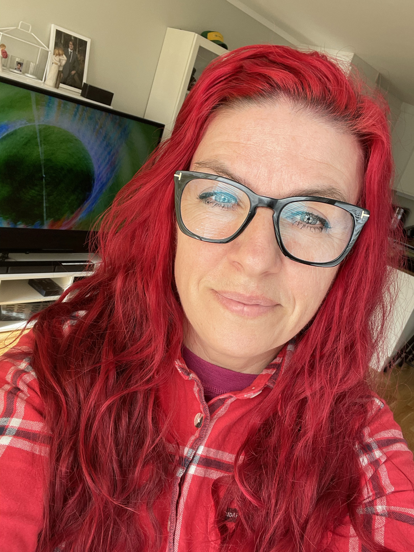 Dame med langt rødt hår, briller og rød, rutete skjorte tar selfie i en stue.