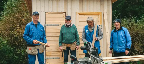 Fire menn i arbeidstøy utenfor en nybygd utedo. Foto.