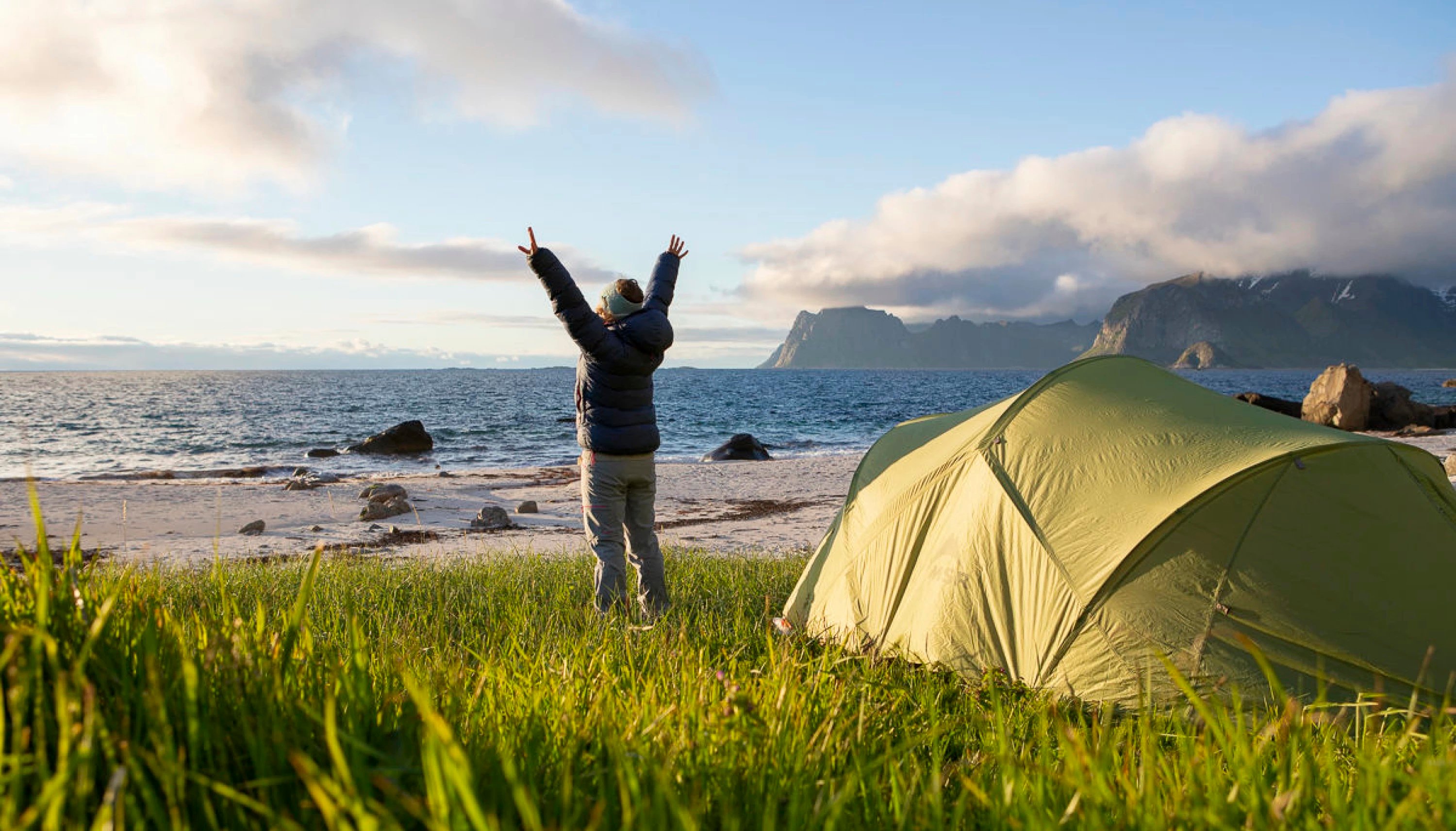 Kvinne i mørkeboble boblejakke står foran et grønt telt på stranda og strekker armene i været.