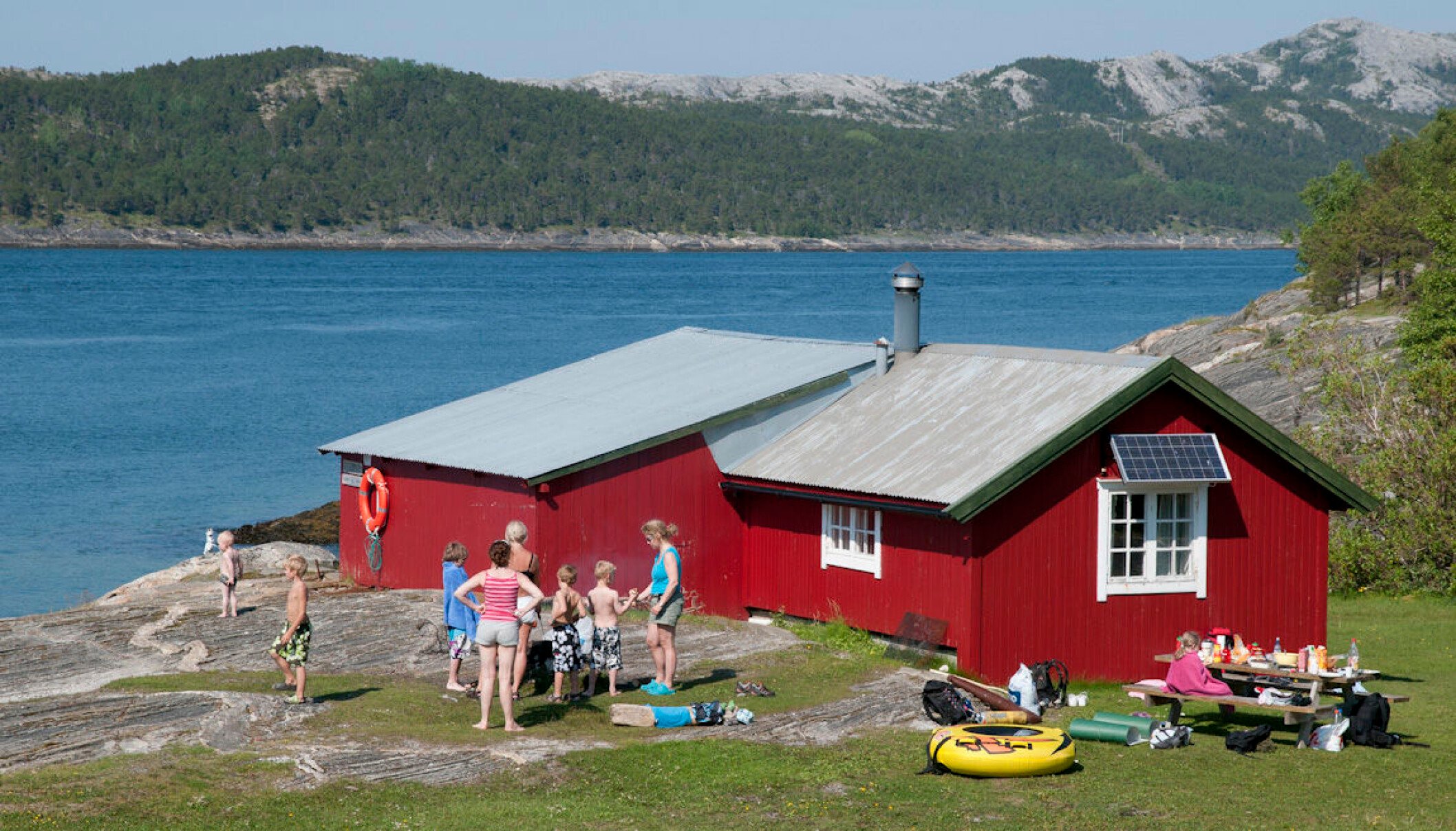 Flere voksne og barn leker og bader utenfor en rød hytte som ligger rett ved sjøen. 