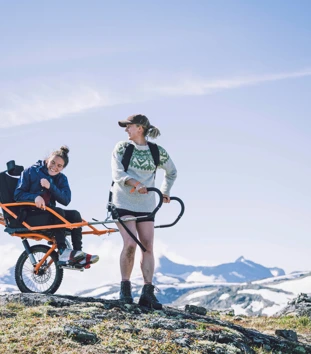 To personer transporterer en tredje person i rullestol med store hjul i fjellet. 