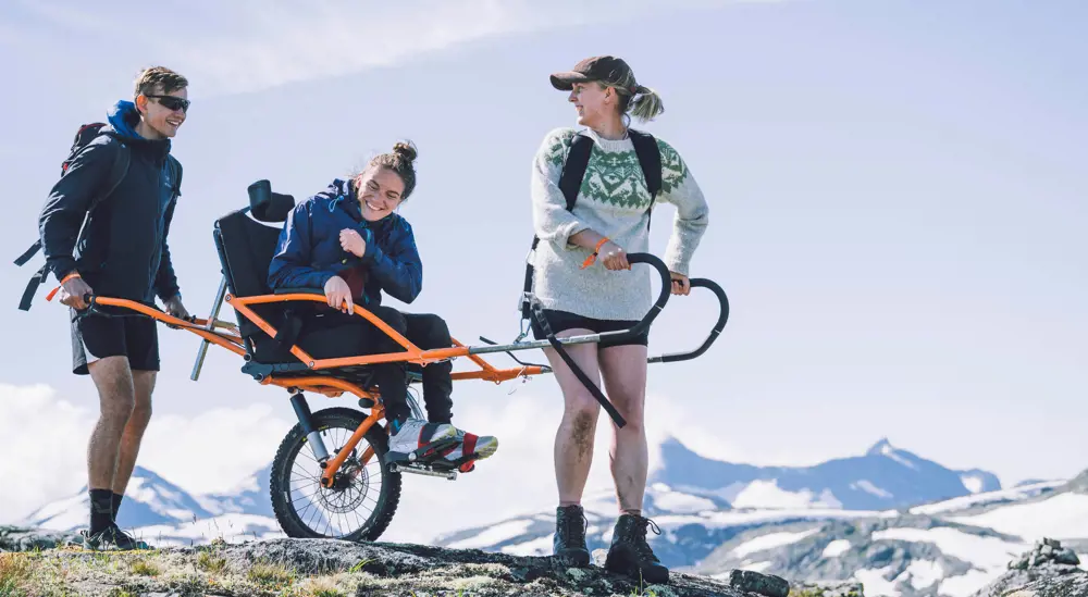 To personer transporterer en tredje person i rullestol med store hjul i fjellet. 