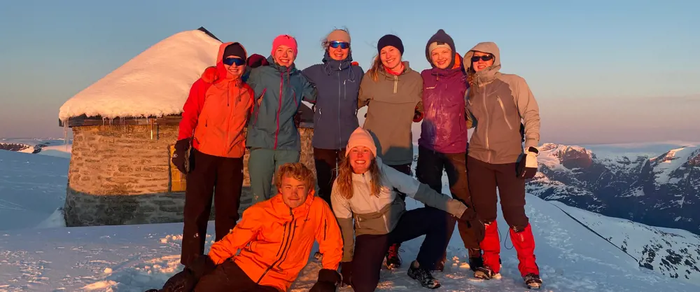 En gruppe glade ungdommer står på Skåla, smiler og ser i kamera. De er vinterkledde og det er snø rundt.