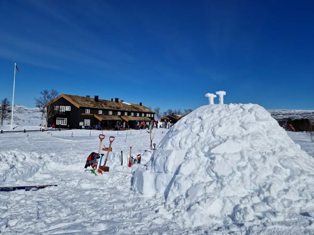 En stor igloo i forgrunnen, med utstyr som spader og sag satt i snøen rundt, og to T-er formet av snø stilt ut på toppen. En lang brun hytte i bakgrunnen, med en gruppe folk vrimlende foran. 