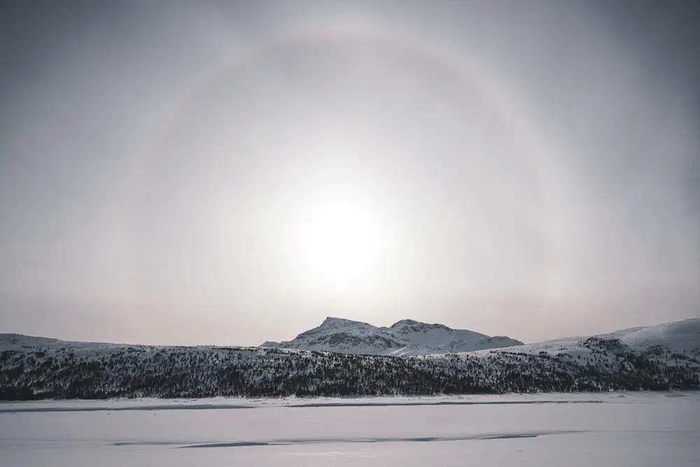  Bildet viser en stor åpen hvit himmel, med en blek sol i midten, omgitt av en ring-halo av lys, med et snødekket fjell rett under, som er del av en fjellkjede som strekker langs horisonten. Snødekket slette i forgrunnen.