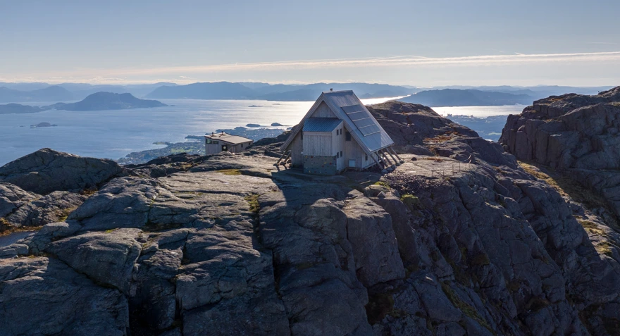 Grå hytte med skrått tak på fjelltopp på Stord med sjø og øyer i bakgrunnen.