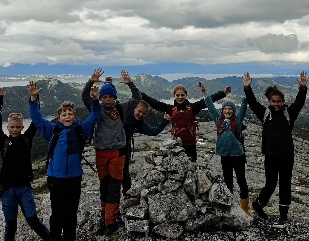 En gjeng ungdommer på toppen av fjellet med hendene opp i været og varden foran seg.