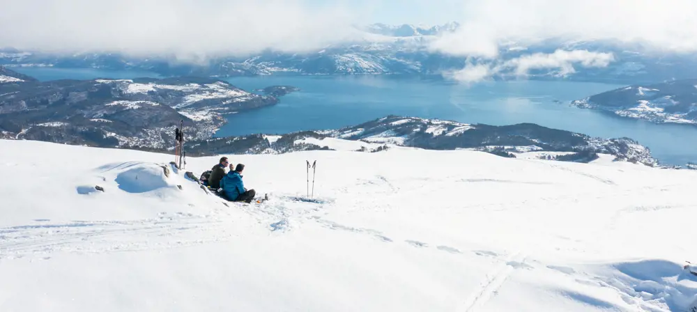To personer har pause i en bakke med snø. Sol og fin utsikt over Hardangerfjorden.