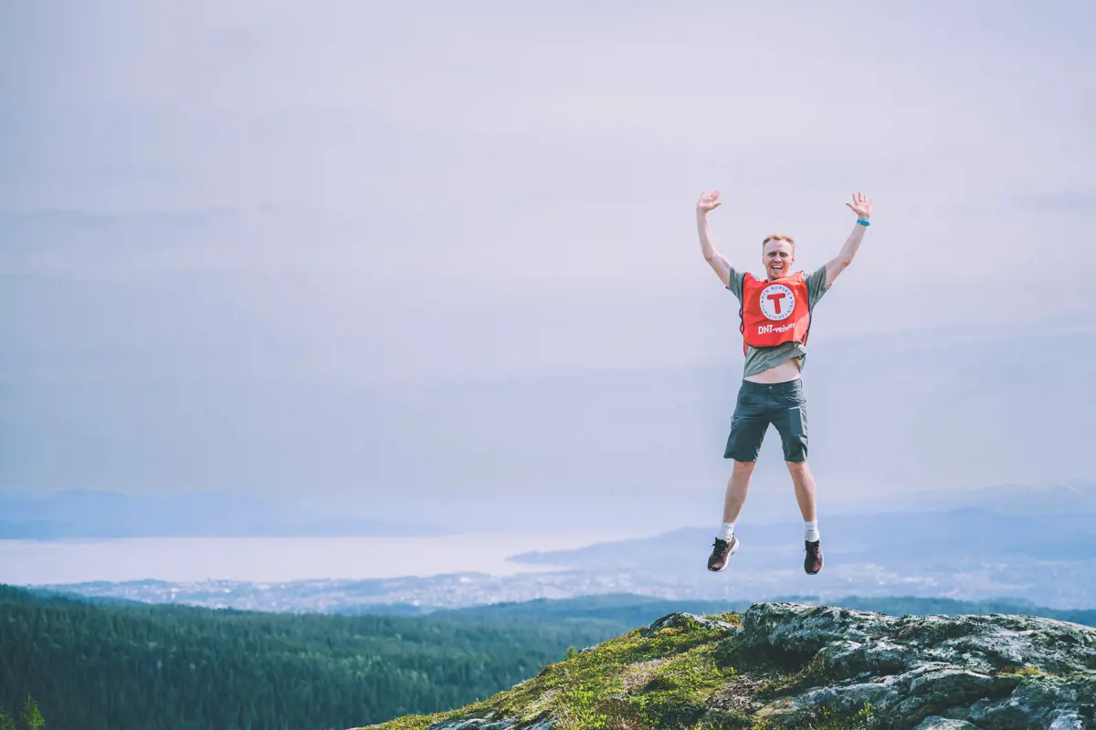 En mann med en rød vest med T for turistforeningen på hopper med armene i været og smiler på en fjelltopp. I bakgrunnen kan man skimte byen Trondheim.