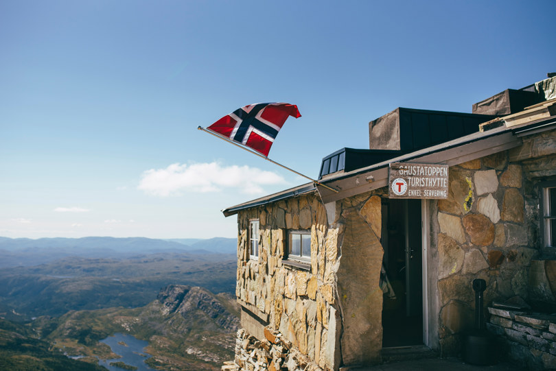Et lite norsk flagg vaier i vinden på veggen av en grå steinhytte. 