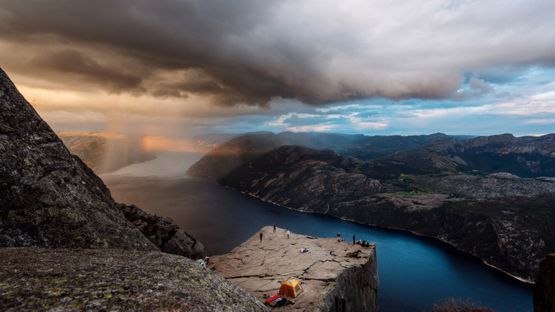Flere mennesker og et gult telt står på en flat fjellhylle over en fjord.