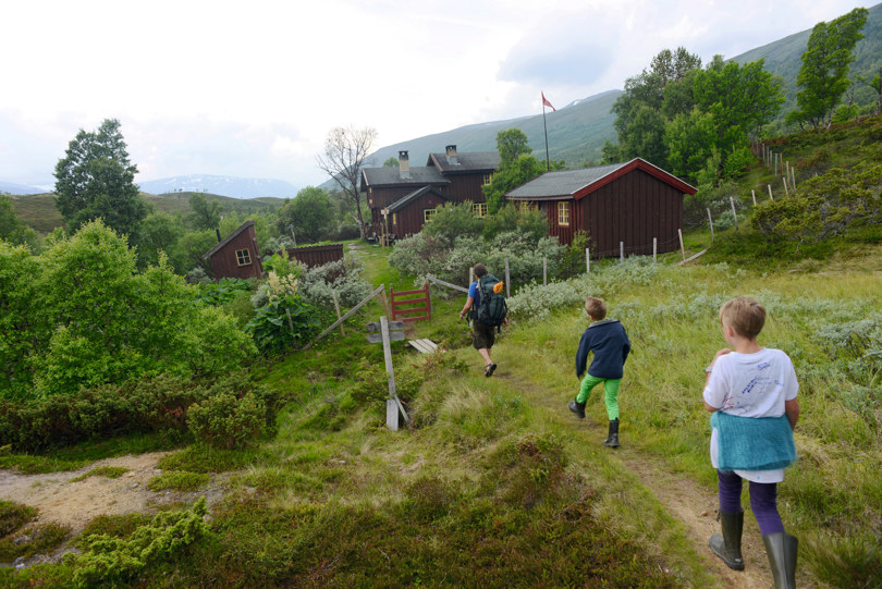 Tre personer går på en sti mot ei hytte i grønt og frodig fjellterreng.