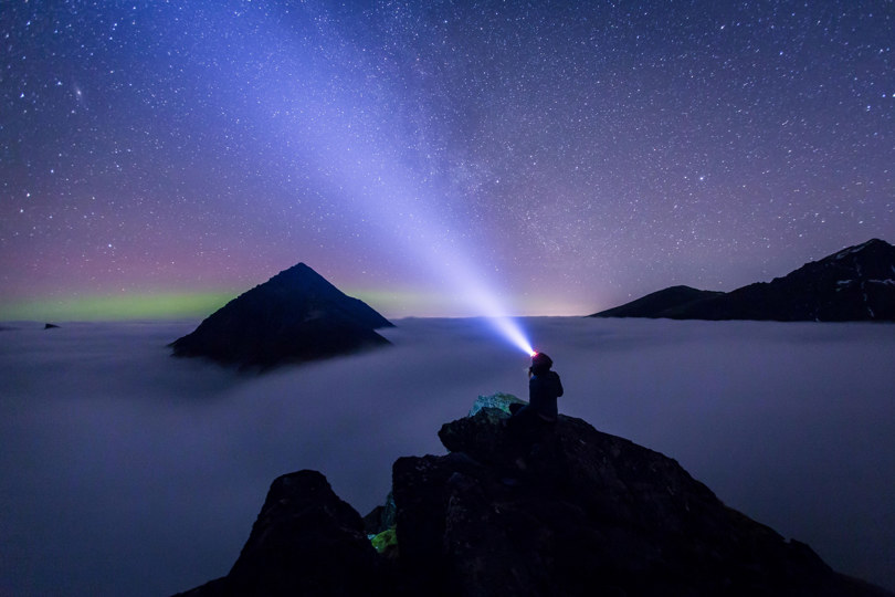 Et menneske sitter i mørket på toppen av et fjell og lyser med hodelykt opp på stjernehimmelen.