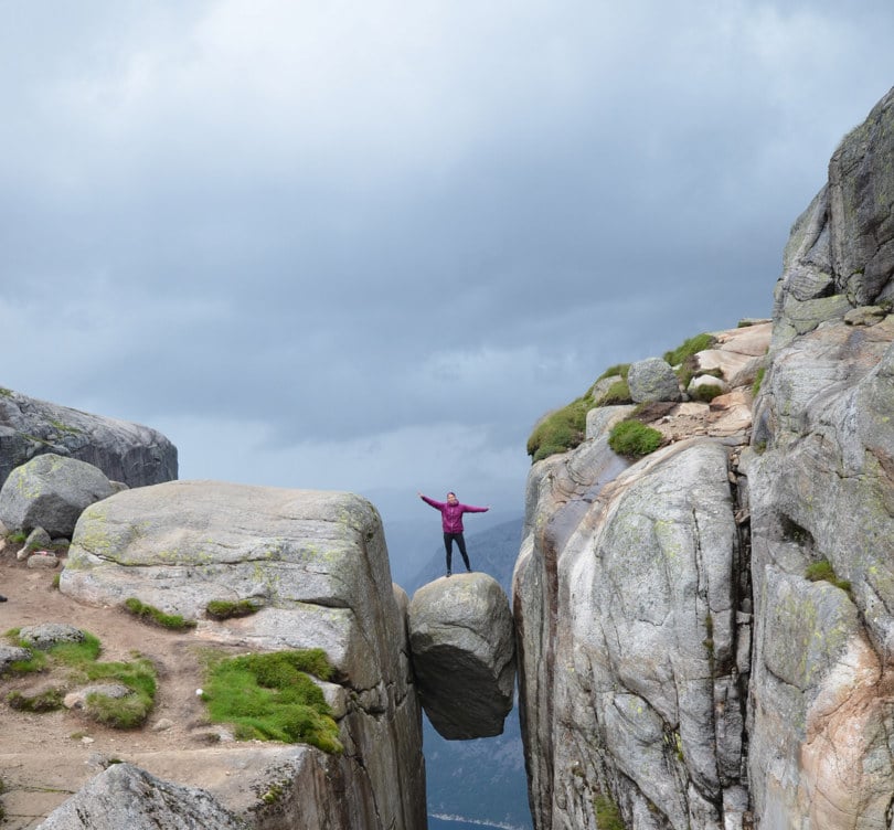 Person i lilla jakke strekker armene i været, stående på en steinblokk som er kilt fast mellom to fjellklipper.