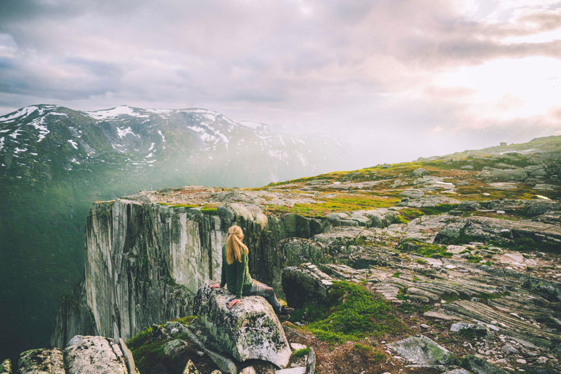 Kvinne i grønn jakke sitter på en stein med ryggen til og ser mot en loddrett fjellside. 