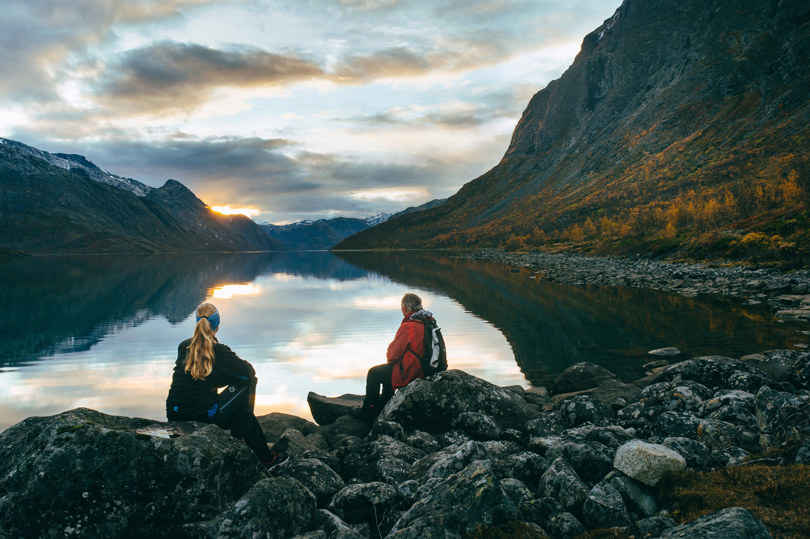 To personer sitter på steiner ved kanten av et fjellvann og ser innover vannet og fjellene rundt.