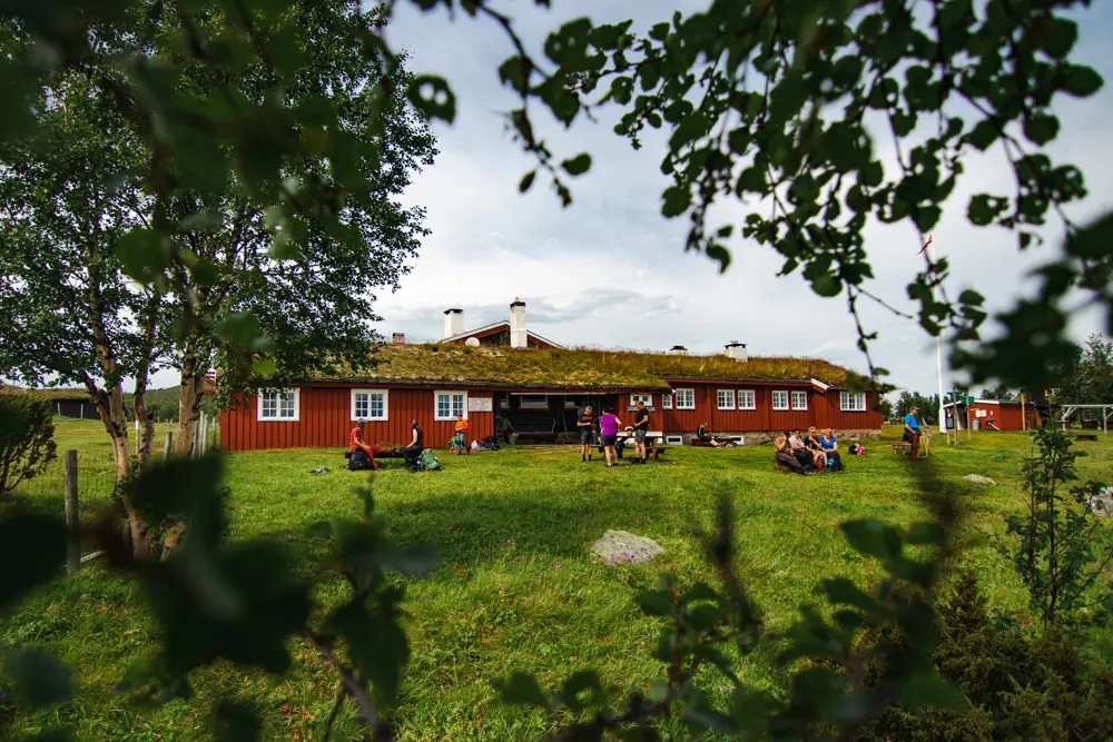 En rød og langstrakt hytte, med et gressdekket landskap foran seg, og noen trær mellom oss og bygningen