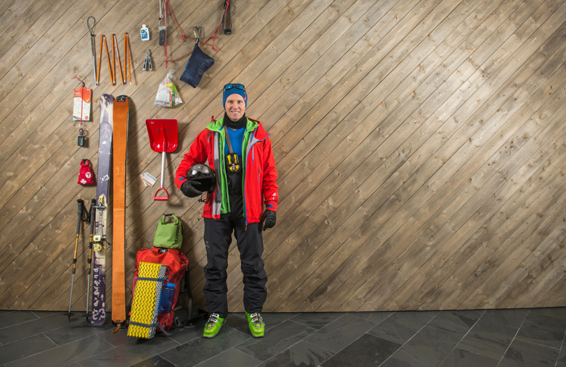 Person ikledd rød skijakke og svart bukse står inntil en vegg med skrå trepanel. Ved siden av henger masse ski- og turutstyr.