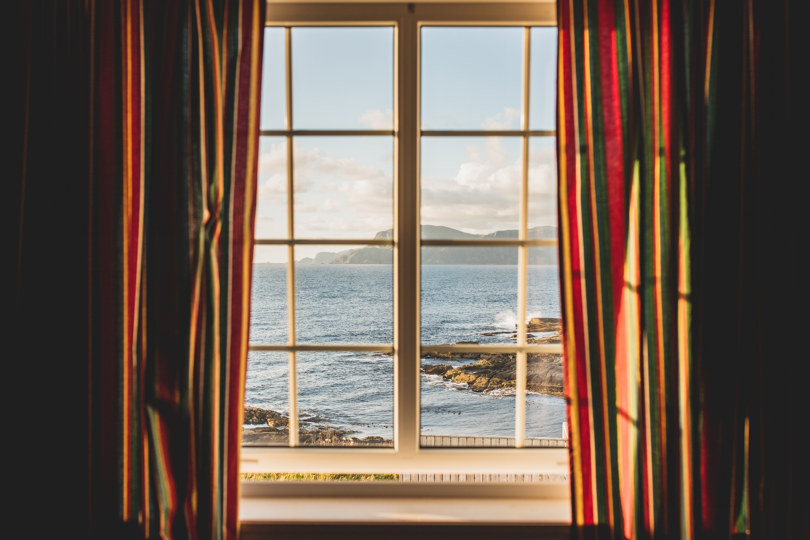 Utsikt mot hav gjennom et vindu med gardiner på hver side.