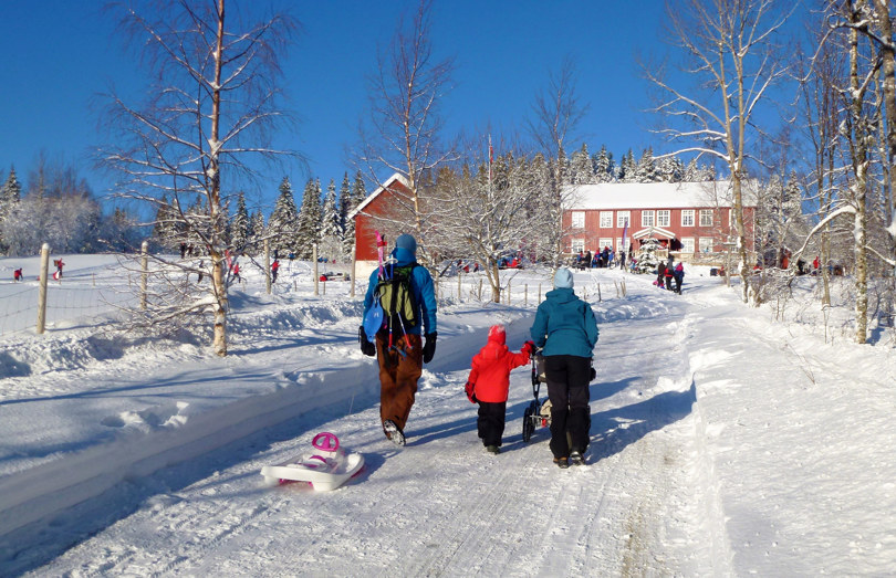 En familie med barn og akebrett går oppover en brøytet vei til en stor rød hytte.