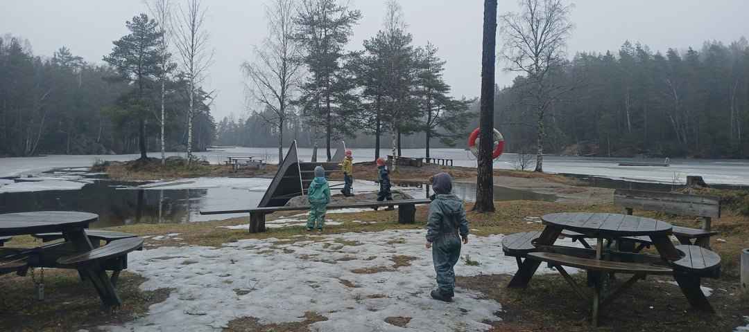 Barn som leker ved et vann. Det er grå himmel, og snøen ligger flekkvis innimellom piknikkbord.