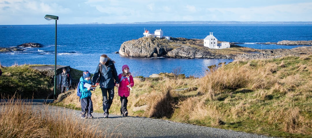 En kvinne med to barn går oppover en grusvei. De holder hender. Bak dem skimtes et fyr og blått hav.