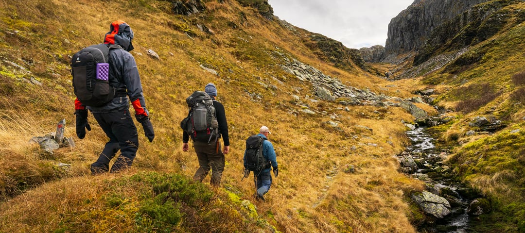 3 personer går tur med sekk. Det er høstfarger som brunt og grått i terrenget. De er godt kledd med votter og hetten oppe. Tydelig at det er vått
