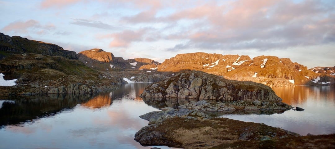 Morgenstemning med soloppgang over et stille vann i fjellet. Det ligger litt snø enkelte steder 