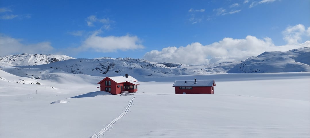 Tomannsbu. 2 røde hytter i snødekt landskap. Det er blå himmel og sol. 