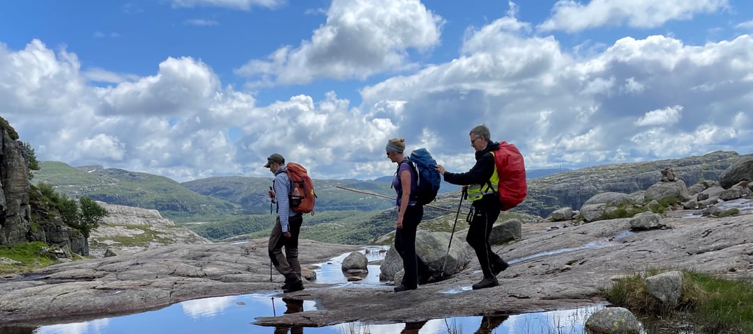 Tre personer går forbi et vann i fjell-landskap. Den bakerste personen på rekka holder i sekken til personen foran og holder en mobilitetsstokk.