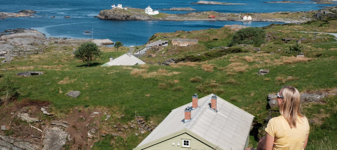 En kvinne sitter i gresset og ser ut over havet. En lysegrønn hytte er nedenfor.