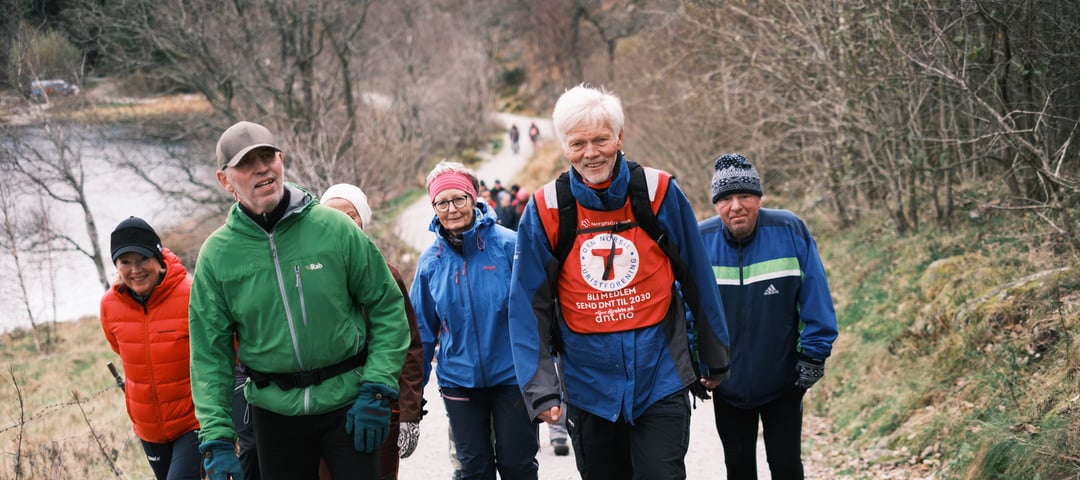 En gruppe turgåere med en turleder med rød T på brystet i front. Alle smiler og går oppover en grusti.
