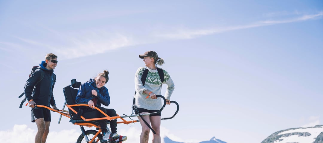 To personer transporterer en tredje person i rullestol med store hjul i fjellet.