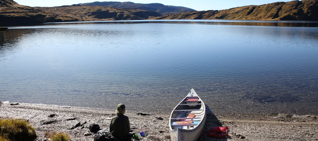 Et barn sitter på steinstrand med kano og ser utover vannet.