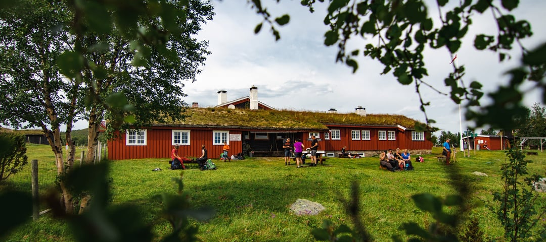 En rød og langstrakt hytte, med et gressdekket landskap foran seg, og noen trær mellom oss og bygningen
