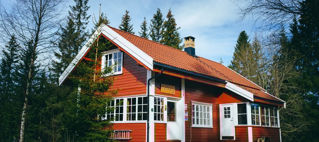 Rødmalt hytte med hvite karmer og oransje takstein i barskog. 