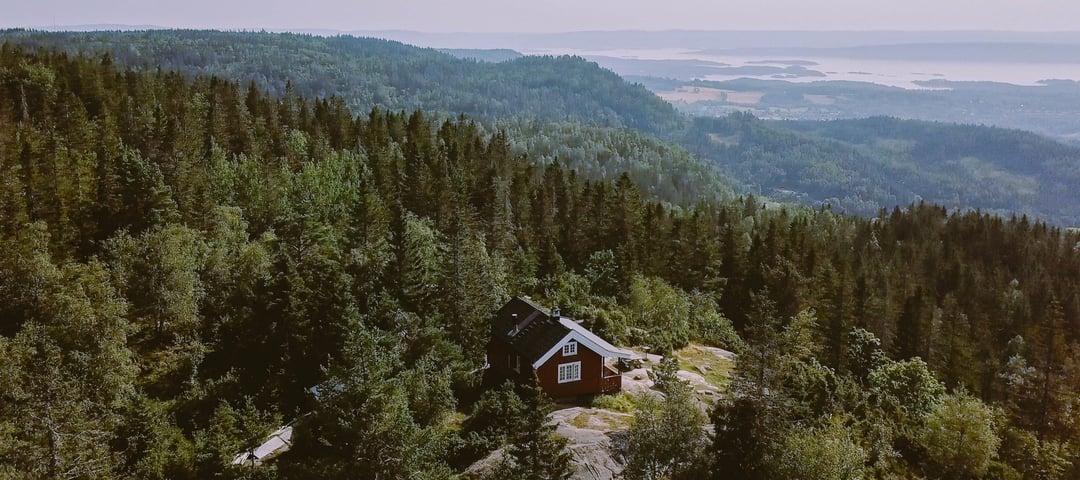 Rød hytte med hvite vinduer omkranset av tett skog og åser.