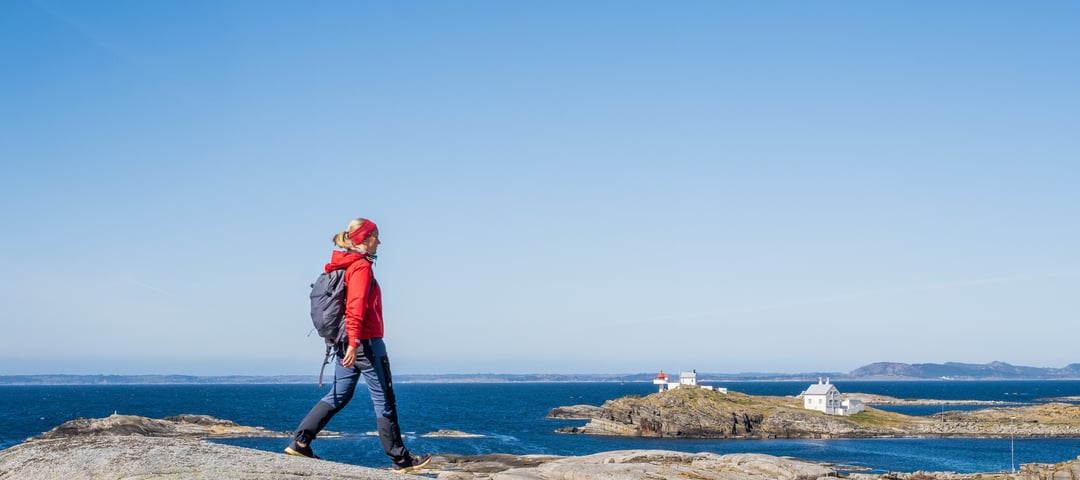 En kvinne i rødt går tur på et berg. Et fyrtårn i vises i bakgrunnen og blått hav