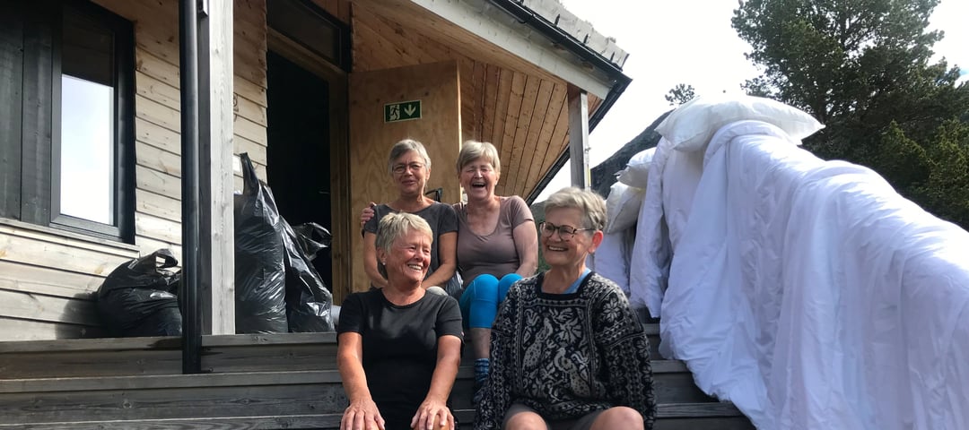 Fire damer sitter på en trapp utenfor en grå hytte. Det henger dyner til lufting.