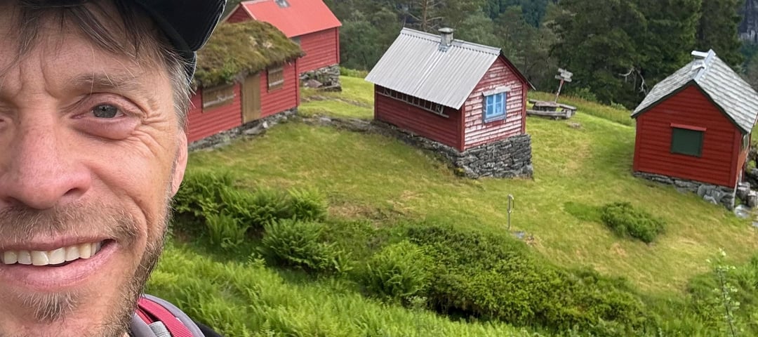 Smilende mann med caps og t-skjorte står foran rødmalt stølstun.