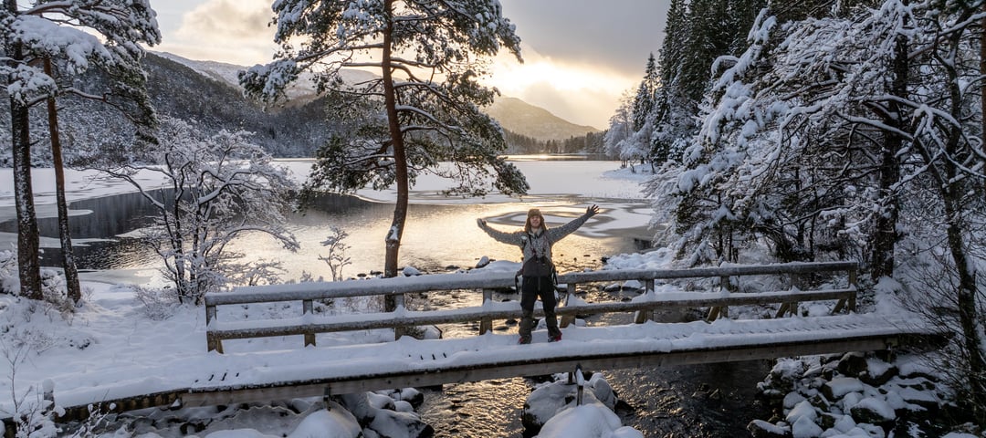 Mann står på bro foran vann og holder armene opp, ønsker velkommen, i vinterlandskap.