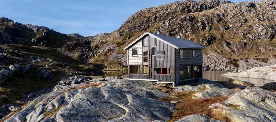 Stor, grå hytte med store vinduer og skråe vinkler i solrikt høstfjell.