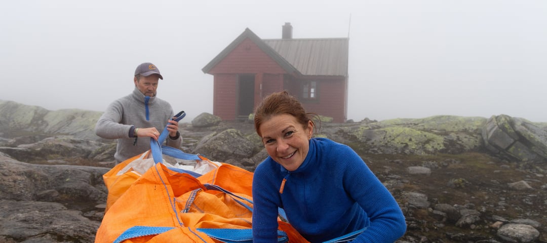 Kvinne i blå genser smiler foran hytte i tåkeheim.