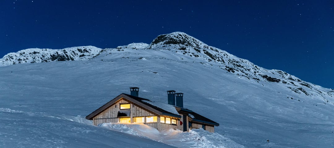 Hytte i vakkert snølandskap i fjellet med lys inne og stjernehimmel over hytta.