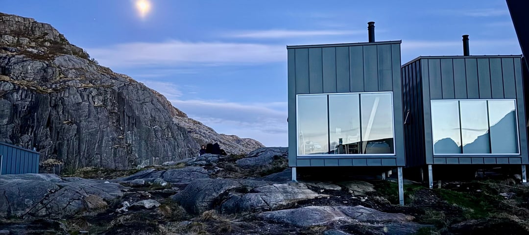 En grå hytte med store vinduer som ligger på fjellet. Måneskinn og kveldslys.