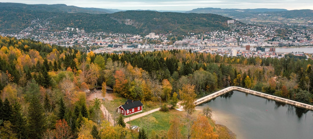 liten rød hytte som ligger i skogen ved et vann. i bakgrunnen ser man utsikt til en større by.